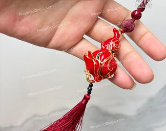 May include: A red gourd-shaped charm with gold trim and black accents, suspended from a red cord with a tassel. The charm is held up by a hand. The charm is a decorative item.