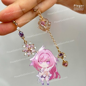 Puede incluir: Un par de pendientes dorados con diseños intrincados. Uno presenta una cadena colgante con un dije en forma de corazón y detalles morados. El otro tiene un diseño floral con cristales rosas y transparentes. Un personaje de dibujos animados está en la imagen.