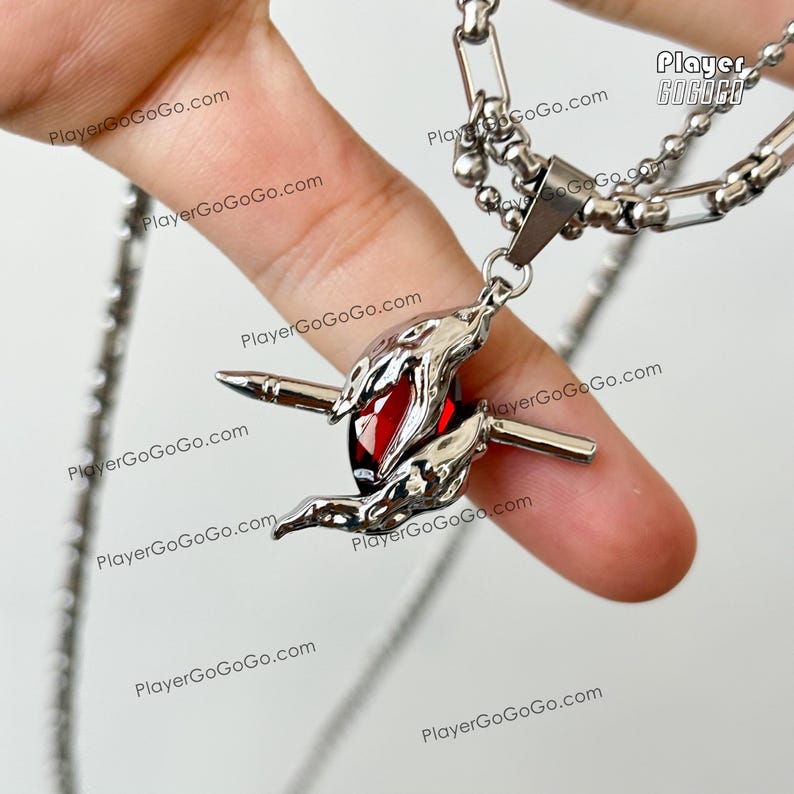 May include: A silver necklace with a red gem pendant. The pendant features two crossed swords with a red gem in the center. The necklace is on a chain with a silver clasp.