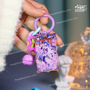 Puede incluir: Un llavero morado con un dije rectangular con una ilustraci&oacute;n de personaje. El dije tiene un borde dorado y un dise&ntilde;o con colores morado, azul y blanco. El llavero incluye un anillo morado, un cierre azul claro y una campana rosa.