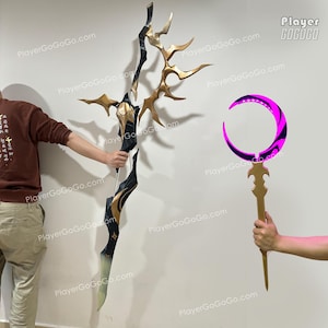 May include: A black and gold bow and a staff with a crescent moon design. The bow has a detailed gold design and is held by a person. The staff has a pink and black crescent moon design and is held by a person. The background is a plain white wall.