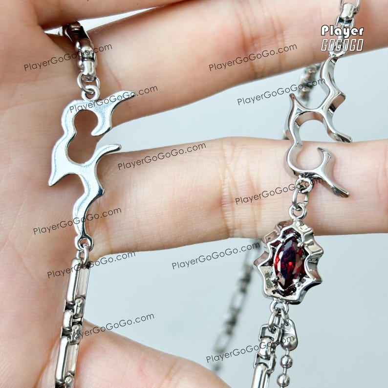May include: Silver chain bracelet with a red gemstone pendant and a silver flame design charm. The bracelet has a double chain design and a lobster clasp.