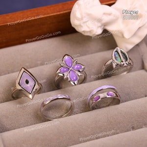 May include: Five silver rings with purple and white enamel designs. The rings are displayed in a velvet-lined box. The rings feature a variety of designs, including a flower, a diamond shape, and a simple band.