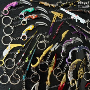 K&ouml;nnte beinhalten: Eine Sammlung bunter Schl&uuml;sselanh&auml;nger mit verschiedenen Waffendesigns, darunter Messer, Schwerter und &Auml;xte. Die Schl&uuml;sselanh&auml;nger sind aus Metall und haben eine silberne Oberfl&auml;che. Die Schl&uuml;sselanh&auml;nger sind alle an silbernen Schl&uuml;sselringen befestigt. Die Schl&uuml;sselanh&auml;nger sind in einem zuf&auml;lligen Muster auf einer schwarzen Oberfl&auml;che angeordnet. Der Text "PlayerGoGoGo.com" erscheint auf mehreren der Schl&uuml;sselanh&auml;nger.