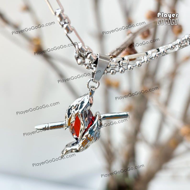 May include: A silver necklace with a pendant featuring a red gemstone surrounded by silver flames. The pendant is attached to a silver chain with a lobster clasp.