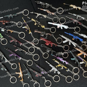 K&ouml;nnte beinhalten: Eine Sammlung von 20 verschiedenen pistolenf&ouml;rmigen Schl&uuml;sselanh&auml;ngern in verschiedenen Farben und Designs. Die Schl&uuml;sselanh&auml;nger sind an silbernen Schl&uuml;sselringen befestigt. Die Schl&uuml;sselanh&auml;nger sind in einem Halbkreis auf einer schwarzen Oberfl&auml;che angeordnet. Der Text "PlayerGOGOGO.com" ist im Hintergrund sichtbar.