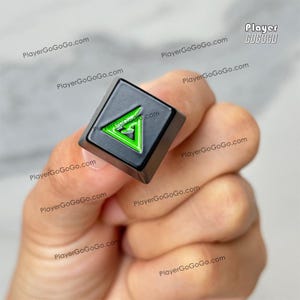 Könnte beinhalten: Eine schwarze quadratische Tastenkappe mit einem neongrünen Dreieck-Design. Auf der Tastenkappe befindet sich der Text "PlayerGoGoGo.com". Die Tastenkappe wird in der Hand einer Person gehalten.