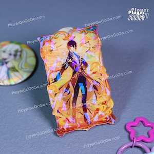 Puede incluir: Un charm acr&iacute;lico hologr&aacute;fico rectangular con una ilustraci&oacute;n de personaje. El charm tiene una combinaci&oacute;n de colores dorados y naranjas con una imagen detallada de una persona con un abrigo largo. El charm est&aacute; rodeado por un borde decorativo.
