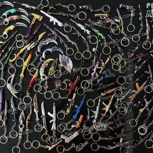 K&ouml;nnte beinhalten: Eine Sammlung bunter Schl&uuml;sselanh&auml;nger mit verschiedenen Waffen und Messern. Die Schl&uuml;sselanh&auml;nger sind aus Metall und haben eine silberne Oberfl&auml;che. Die Schl&uuml;sselanh&auml;nger sind in einem zuf&auml;lligen Muster auf einer schwarzen Oberfl&auml;che angeordnet. Der Text "Player GoGoGo.com" ist im Bild sichtbar.