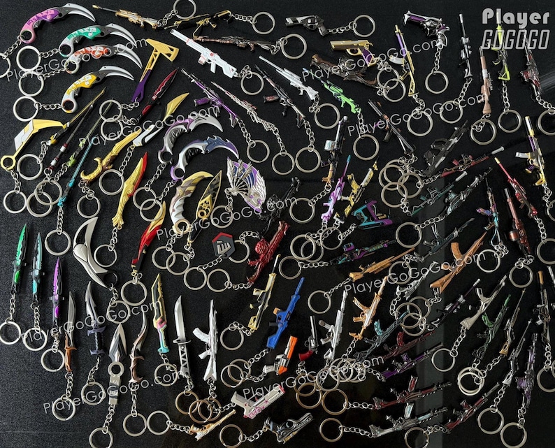 K&ouml;nnte beinhalten: Eine Sammlung bunter Schl&uuml;sselanh&auml;nger mit verschiedenen Waffen und Messern. Die Schl&uuml;sselanh&auml;nger sind aus Metall und haben eine silberne Oberfl&auml;che. Die Schl&uuml;sselanh&auml;nger sind in einem zuf&auml;lligen Muster auf einer schwarzen Oberfl&auml;che angeordnet. Der Text "Player GoGoGo.com" ist im Bild sichtbar.
