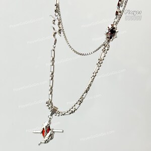 May include: A silver chain necklace with two pendants. The top pendant is a silver star with a red center. The bottom pendant is a silver swirl with a red center. The necklace is on a white background.