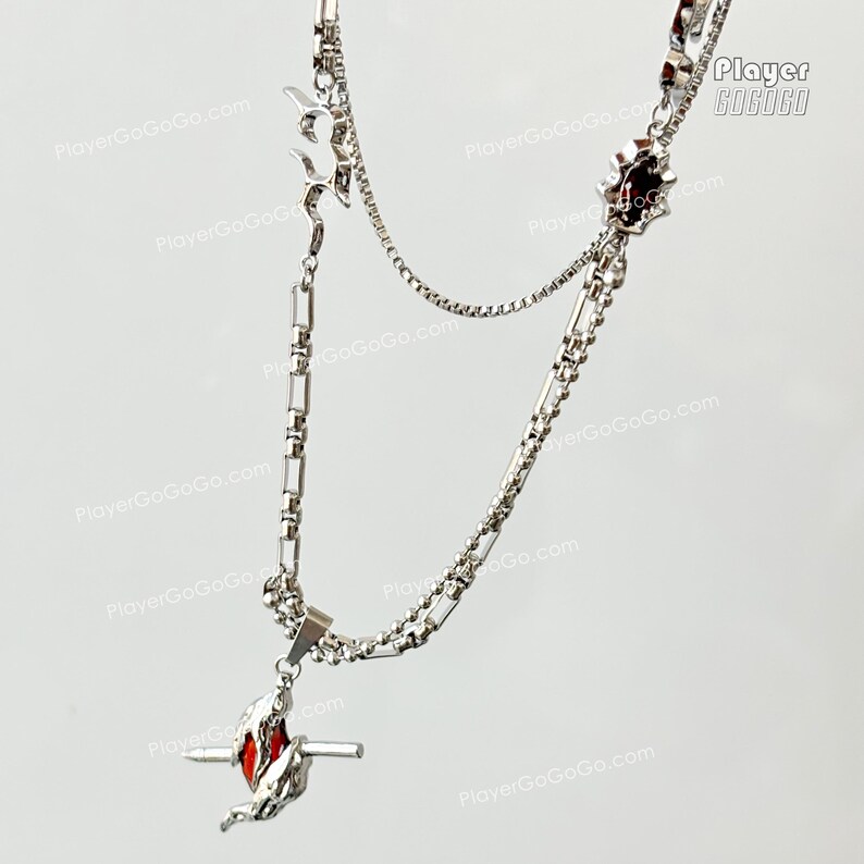May include: A silver chain necklace with two pendants. The top pendant is a silver star with a red center. The bottom pendant is a silver swirl with a red center. The necklace is on a white background.
