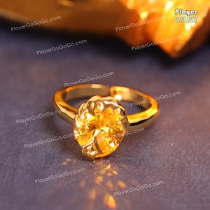 May include: A gold ring with a large, round, yellow gemstone. The ring is set on a dark gray surface.
