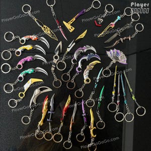 K&ouml;nnte beinhalten: Eine Sammlung von 20 bunten Schl&uuml;sselanh&auml;ngern aus Metall mit verschiedenen Waffendesigns, darunter Messer, Schwerter und &Auml;xte. Die Schl&uuml;sselanh&auml;nger sind auf einem schwarzen Hintergrund angeordnet, auf dem der Text "PlayerGoGoGo.com" zu sehen ist.