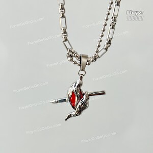 May include: A silver chain necklace with a pendant featuring a red gem surrounded by two silver wings and a silver spear. The pendant is in the shape of a stylized bird.