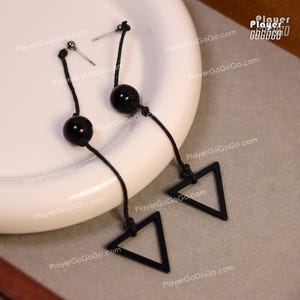 Puede incluir: Pendientes colgantes negros con un diseño minimalista. Cada pendiente presenta una cuenta negra suspendida de un poste, conectada a un triángulo negro. Los pendientes se exhiben en un plato blanco.