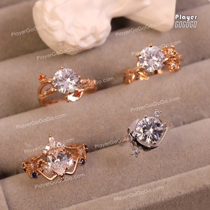 Puede incluir: Cuatro anillos de oro con piedras preciosas transparentes y diseños intrincados. Los anillos se muestran en una superficie beige. PlayerGoGoGo.com está impreso en la superficie.