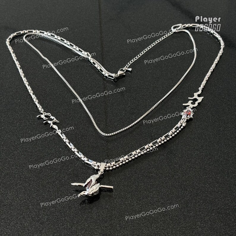 May include: A silver layered necklace with a red stone pendant. The pendant features a stylized design with a wave-like pattern. The necklace is made of three different chain styles.