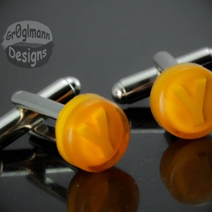 May include: A pair of silver cufflinks with amber-colored round faceted glass inserts. Each insert features a raised letter "V" in the center.