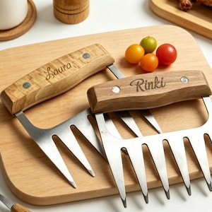 May include: Two stainless steel meat shredder claws with wooden handles. One handle is engraved with the name Laura, and the other is engraved with the name Rinki. Each tool features four sharp, pointed metal prongs for shredding cooked meat.