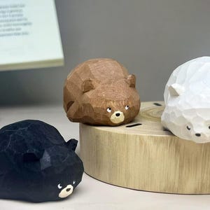May include: Three small, hand-carved wooden bear figurines in black, brown, and white. Each bear features a faceted, geometric texture, small rounded ears, and a painted face with white eyes and a snout. The figures are arranged together.