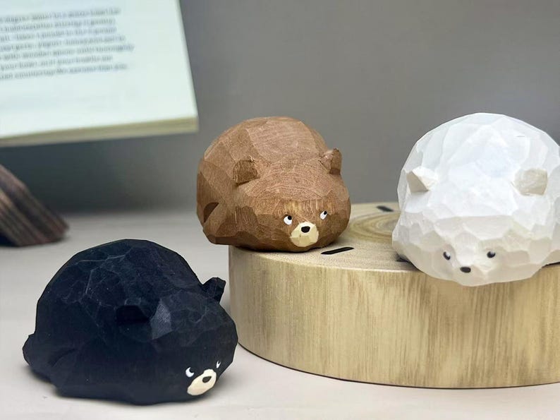 May include: Three small, hand-carved wooden bear figurines in black, brown, and white. Each bear features a faceted, geometric texture, small rounded ears, and a painted face with white eyes and a snout. The figures are arranged together.