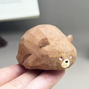 May include: A small, hand-carved wooden bear figurine with a faceted, textured surface. The bear is light brown with a lighter cream-colored muzzle, a black nose, and two small, dark, forward-facing eyes. The sculpture has rounded ears.