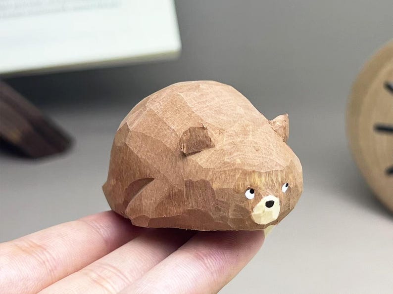 May include: A small, hand-carved wooden bear figurine with a faceted, textured surface. The bear is light brown with a lighter cream-colored muzzle, a black nose, and two small, dark, forward-facing eyes. The sculpture has rounded ears.