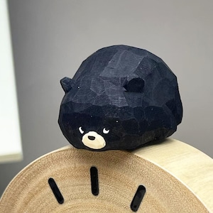 May include: A small, hand-carved wooden bear figurine with a matte black finish, featuring a rounded body, two small ears, and a white muzzle with black eyes. The bear is positioned on top of a light-colored, circular wooden clock face with black markers.