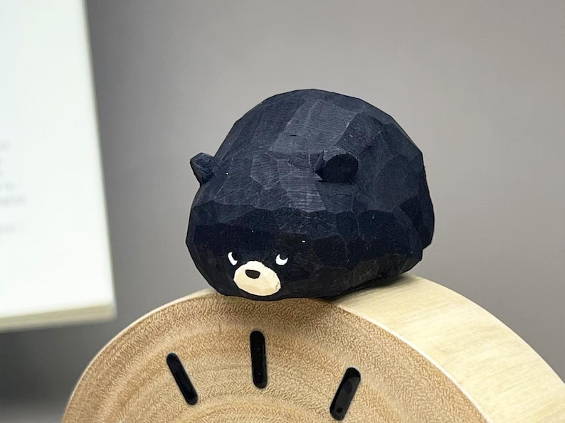 May include: A small, hand-carved wooden bear figurine with a matte black finish, featuring a rounded body, two small ears, and a white muzzle with black eyes. The bear is positioned on top of a light-colored, circular wooden clock face with black markers.