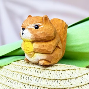 May include: A hand-carved wooden squirrel figurine. The squirrel is light brown with a yellow belly and a bushy tail. It has black eyes and is sitting on a woven surface with a green leaf in the background.