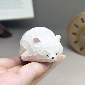 May include: A small, white, hand-carved wooden figurine of a bear with a faceted, geometric surface texture. The bear has two small ears, a rounded body, and painted black eyes and a nose, presented in a compact, stylized, and smooth form.