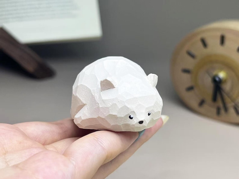 May include: A small, white, hand-carved wooden figurine of a bear with a faceted, geometric surface texture. The bear has two small ears, a rounded body, and painted black eyes and a nose, presented in a compact, stylized, and smooth form.