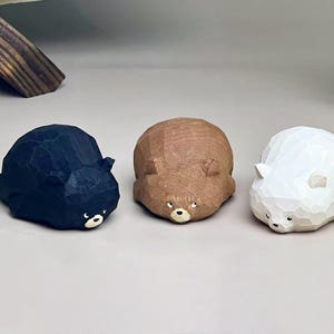 May include: Three small, faceted animal figurines carved from wood. The set includes one black bear, one brown bear, and one white bear. Each figurine features a low-profile, rounded shape with visible carved textures, small ears, and painted eyes and noses.