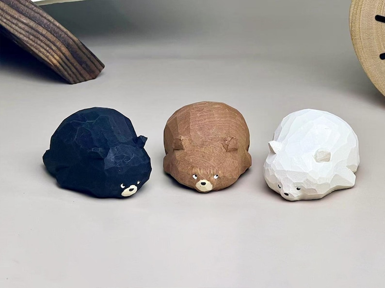 May include: Three small, faceted animal figurines carved from wood. The set includes one black bear, one brown bear, and one white bear. Each figurine features a low-profile, rounded shape with visible carved textures, small ears, and painted eyes and noses.