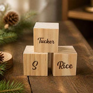 May include: Three light-colored wooden cubes arranged in a pyramid formation. The top cube features the word Tucker in dark brown script. The bottom left cube displays the letter S, and the bottom right cube features the word Rice in dark brown script.