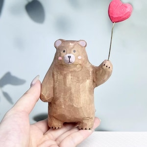 Peut inclure: Une petite figurine d'ours en bois sculpté à la main avec une finition brun clair. L'ours a des joues roses peintes, un nez noir et de petites oreilles. Il tient un fil métallique fin surmonté d'un cœur rouge sculpté dans sa patte gauche.