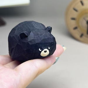 May include: A small, hand-carved wooden figurine of a black bear with a rounded, faceted body. The bear features a light tan muzzle, a black nose, and two small ears. The surface shows visible carving marks, giving it a textured, matte appearance.