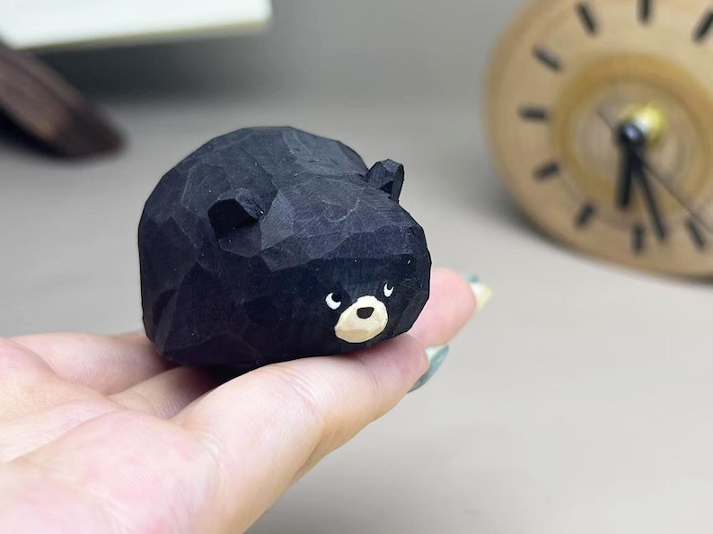 May include: A small, hand-carved wooden figurine of a black bear with a rounded, faceted body. The bear features a light tan muzzle, a black nose, and two small ears. The surface shows visible carving marks, giving it a textured, matte appearance.