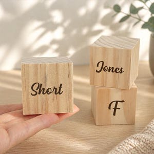 May include: Three light-colored wooden blocks with visible grain patterns. One block is held, featuring the word Short in black script. Two stacked blocks sit nearby, with the top one displaying the word Jones and the bottom one showing the letter F.