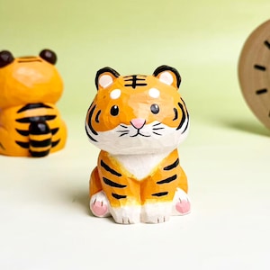 Può includere: Una piccola statuetta di tigre intagliata a mano in legno, caratterizzata da un manto arancione brillante con strisce nere, petto bianco e naso rosa. La tigre è raffigurata in posizione seduta con texture intagliate visibili e orecchie arrotondate.
