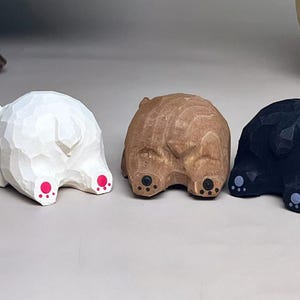 May include: Three small hand-carved wooden animal figurines shown from the rear. One is painted white with bright pink paw pads, one is natural light brown wood with black paw pads, and one is painted black with light gray paw pads.