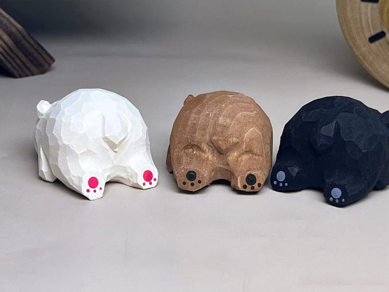 May include: Three small hand-carved wooden animal figurines shown from the rear. One is painted white with bright pink paw pads, one is natural light brown wood with black paw pads, and one is painted black with light gray paw pads.