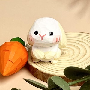 Peut inclure: Une figurine de lapin en bois sculpté à la main avec une finition peinte blanche et jaune. Le lapin a des joues roses et des yeux noirs. Le lapin est assis sur une base en bois avec une carotte à côté.