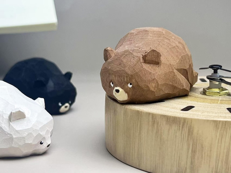 May include: A brown carved wooden bear figurine with faceted surfaces, small ears, and a light-colored snout, positioned on a circular wooden base with a clock mechanism. Two additional bear figurines in white and black are visible nearby.