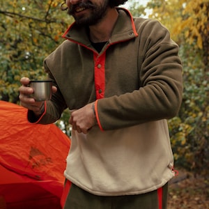 Puede incluir: Jersey de forro polar verde oliva y beige con ribete naranja y pantalón verde oliva a juego. El jersey tiene cuello abotonado. Se sostiene una taza de acero inoxidable en la mano. La escena está ambientada al aire libre con una tienda de campaña al fondo.