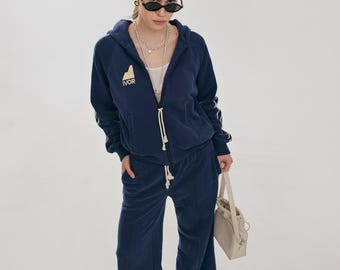 May include: A navy blue tracksuit with a zip-up hoodie and matching wide-leg pants. The hoodie features a white drawstring and the word "IVOR" in gold lettering. The outfit is paired with a cream-colored handbag and sunglasses.