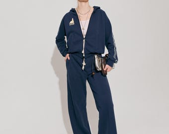Puede incluir: Chándal azul marino con chaqueta con capucha y pantalón de pernera ancha. La chaqueta tiene rayas blancas en las mangas y la palabra "IVOR". La persona lleva gafas de sol y un pequeño bolso plateado. Estilo casual.