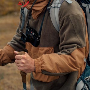 Może przedstawiać: Osoba w brązowej i zielonej kurtce polarowej z brązowo-beżową warstwą zewnętrzną. Niesie duży plecak i kij trekkingowy z nadrukiem "KOMPERDELL".