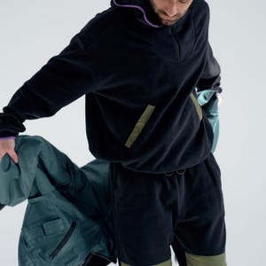 May include: Black fleece hoodie and matching sweatpants with olive green accents. The hoodie has a purple trim and a front pocket. The sweatpants have olive green knee patches. A teal beanie completes the outfit. The person is holding a teal jacket.